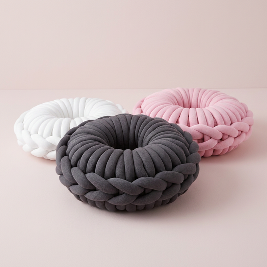 Three round braided donut pilliws  in white, dark gray, and light pink, arranged on a soft blush pink studio background. Made of chunky cotton yarn