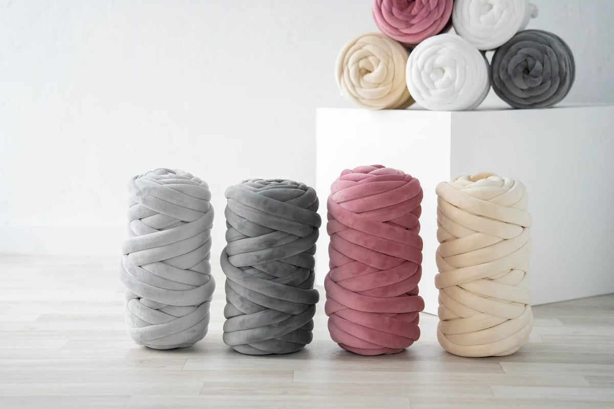 Four thick braided rope cords in light gray, charcoal gray, dusty pink, and cream beige, displayed on a light wood floor with additional rolls stacked on a white block in the background.
