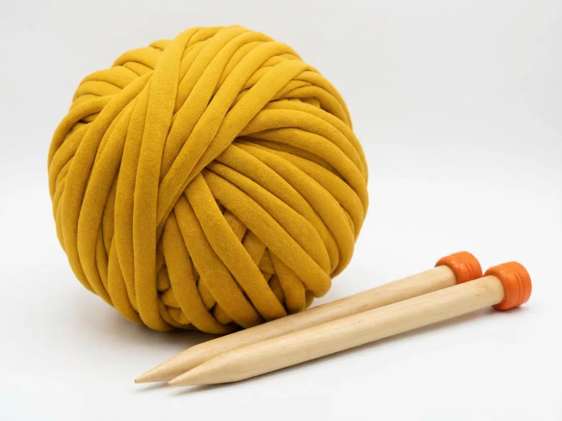 A large ball of thick chunky cotton mustard yarn with two wooden knitting needles with orange end caps, set against a plain white background.