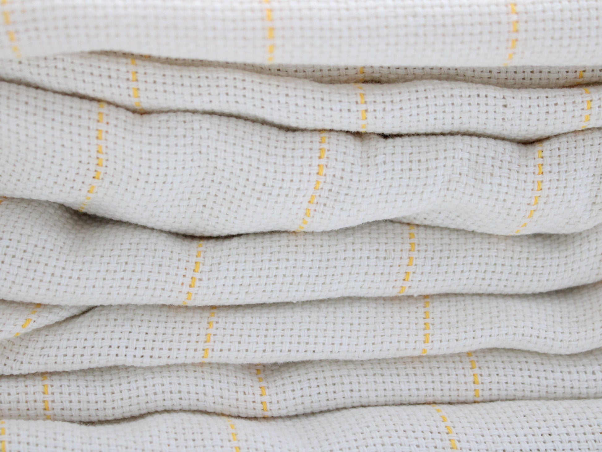 A close-up of folded tufting fabric, cotton  in a warm off-white or cream color with thin vertical yellow stripes.