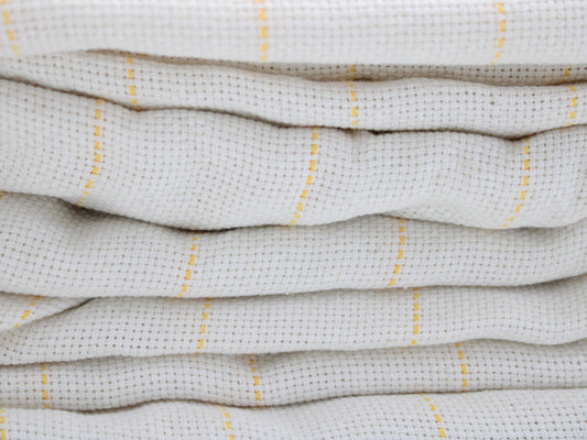 A close-up of folded tufting fabric, cotton  in a warm off-white or cream color with thin vertical yellow stripes.