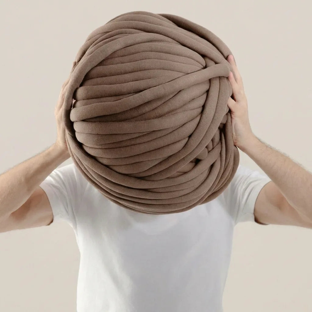Person holding a large ball of taupe cotton tube yarn against a beige background