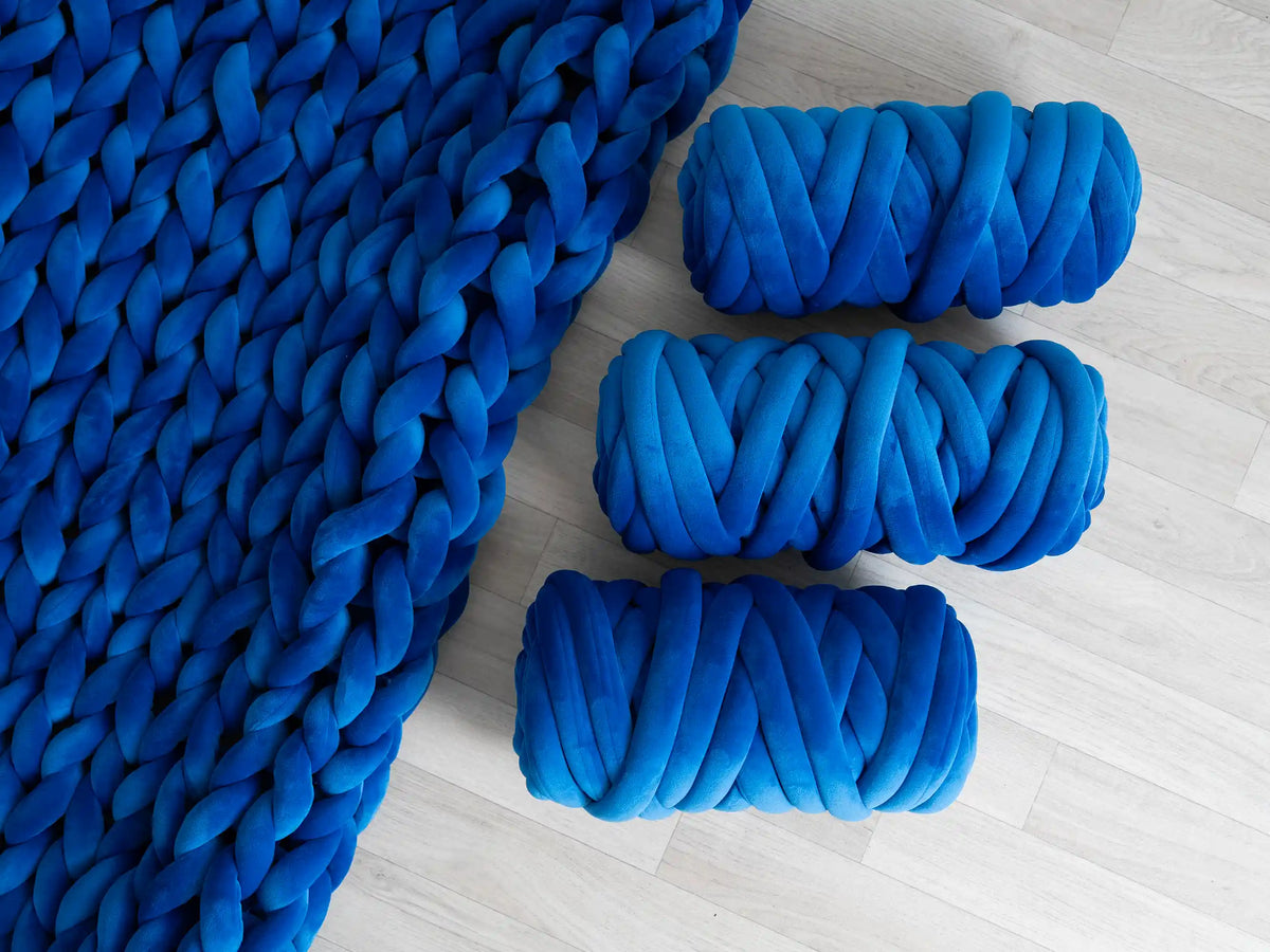 A vibrant blue chunky knit blanket with thick braided loops, shown alongside three matching rolled-up blue cushions on a light gray wood floor.