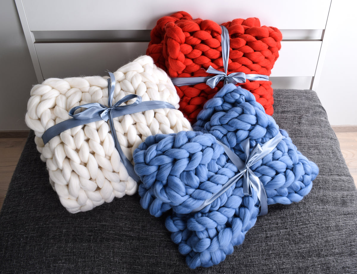 Three chunky merino wool knit throw blankets in white, blue, and red, each tied with a light blue ribbon, arranged on a dark gray cushion. 