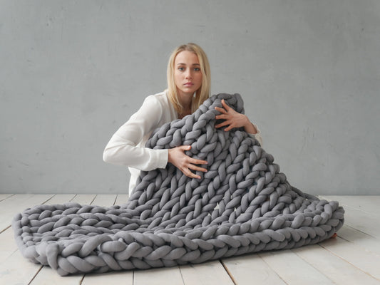 A woman sits on a light wood floor against a gray wall, holding a large chunky knit blanket in a very light gray color. The blanket is spread out in front of her, showing its thick braided texture.