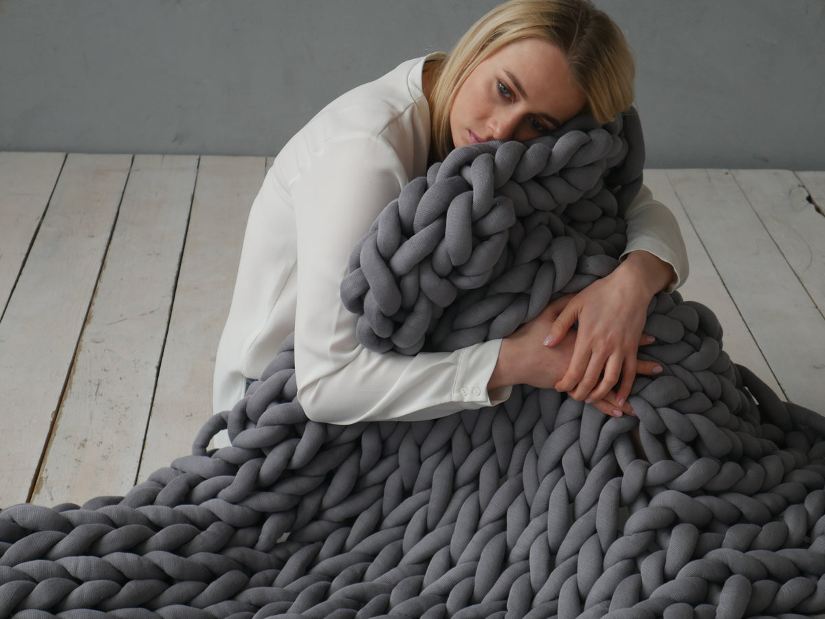 A woman in a white long-sleeve shirt rests her head on a thick, chunky knitted gray weighted blanket, which is spread across a light wood plank floor against a gray wall.