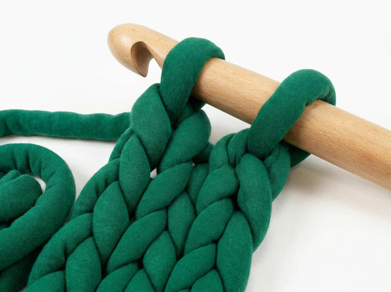 Close-up of a thick green tube yarn or wool chunky cable knit pattern wrapped around a wooden crochet hook on a white background.