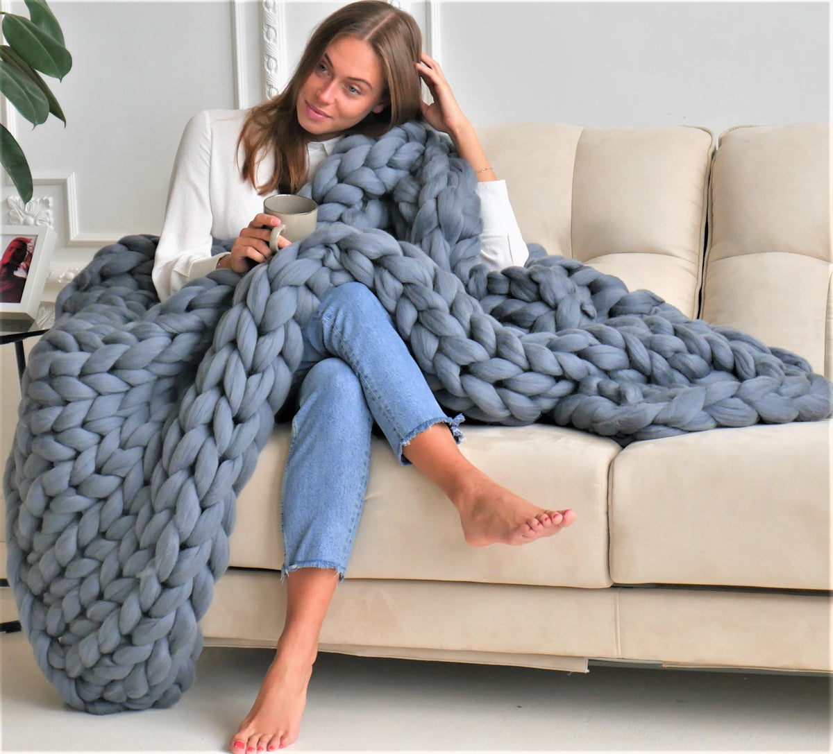 A woman sits on a cream-colored sofa wrapped in a thick, chunky knitted merino wool blue-gray blanket. She is wearing a white long-sleeve top and light blue jeans, holding a mug.